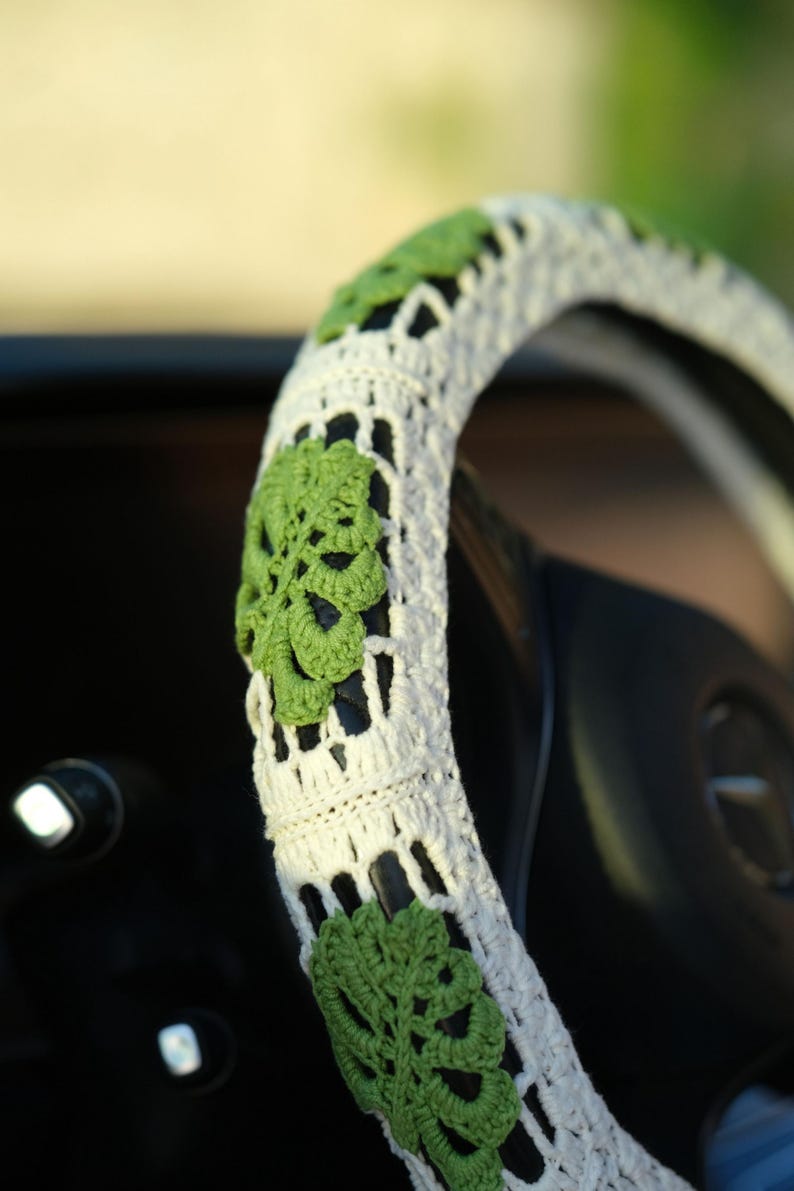 Close-up of a white crochet steering wheel cover decorated with textured green Monstera leaves.