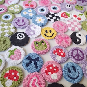 May include: A collection of colorful, round, embroidered patches with various designs, including a smiley face, a mushroom, a peace sign, a frog, a yin yang symbol, a strawberry, a bow, a checkered pattern, a daisy, an eye, a 8-ball, and a cherry.