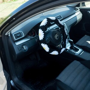 K&ouml;nnte beinhalten: Schwarzes Autoinnenraum mit einer wei&szlig;en und gelben G&auml;nsebl&uuml;mchen-Lenkradabdeckung. Das Lenkrad hat eine schwarze Lederabdeckung und ein silbernes Volkswagen-Logo.