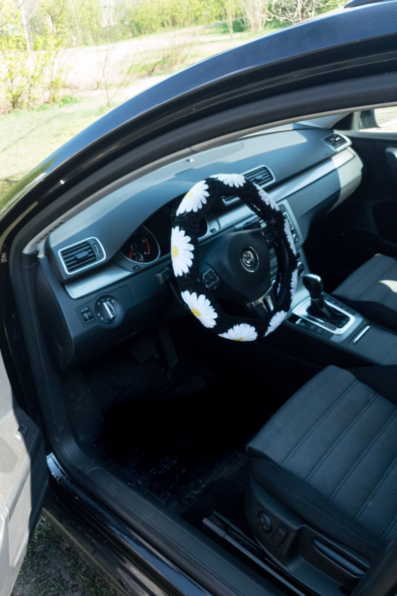 K&ouml;nnte beinhalten: Schwarzes Autoinnenraum mit einer wei&szlig;en und gelben G&auml;nsebl&uuml;mchen-Lenkradabdeckung. Das Lenkrad hat eine schwarze Lederabdeckung und ein silbernes Volkswagen-Logo.