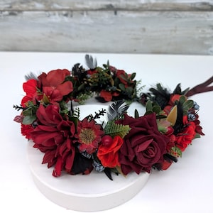May include: A floral crown featuring deep red and burgundy flowers, accented with black feathers and greenery. The crown is arranged on a white surface, showcasing the vibrant colors and textures of the blooms. A dark ribbon is attached.