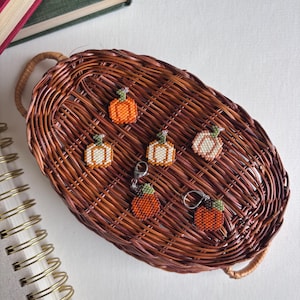 Puede incluir: Pendientes y abalorios de calabaza con cuentas en tonos naranja, blanco y verde, expuestos sobre una bandeja de mimbre marrón tejido. Los pendientes y abalorios están detallados con pequeñas cuentas, sobre una superficie blanca.