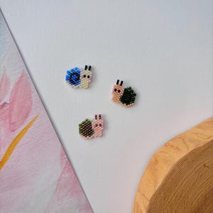 Puede incluir: Tres pequeños abalorios de caracol de cuentas, uno azul, uno verde y uno rosa. Los abalorios están sobre un fondo blanco.