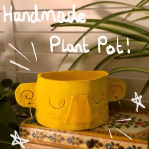 May include: Handmade yellow ceramic plant pot with a face, closed eyes, and a nose. The pot is sitting on a wooden surface with a floral pattern.