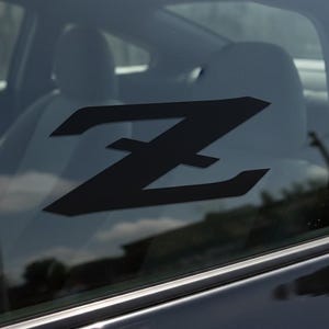 Puede incluir: Calcomanía negra con el logotipo "Z" aplicada a la ventana de un coche. La letra estilizada es un diseño geométrico audaz. El interior del coche es visible a través de la ventana, con los asientos y reposacabezas al fondo.