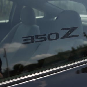 May include: A close-up of a car window with the black text "350Z" visible. The interior of the car is visible, including the seats. The car's exterior is dark, and the window reflects the sky.