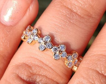 May include: A gold-toned ring featuring a floral design. Each flower is adorned with small, clear gemstones. The ring is shown on a finger, with a blurred green background.