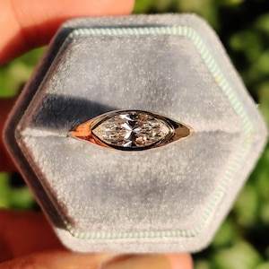Puede incluir: Un anillo de oro con un diamante talla marquesa engastado en un bisel. El anillo se muestra en una caja de anillo de terciopelo azul claro. El anillo es sostenido por una mano contra un fondo verde.