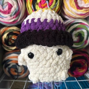 May include: A small, plush, crocheted ghost figure featuring a white body with two black plastic eyes. The ghost wears a layered hat with a yellow top, a purple middle band, and a thick black brim, all crafted from soft, textured yarn material.