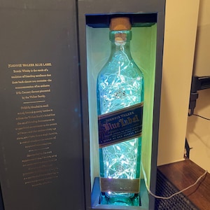 May include: A clear glass bottle of Johnnie Walker Blue Label Scotch Whisky with a blue and white label. The bottle is illuminated with white fairy lights and is sitting in a black box.