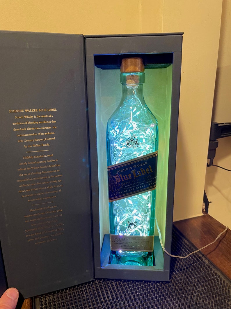 May include: A clear glass bottle of Johnnie Walker Blue Label Scotch Whisky with a blue and white label. The bottle is illuminated with white fairy lights and is sitting in a black box.