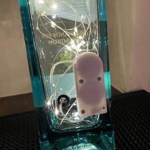 May include: A clear glass bottle with a blue label and a barcode. The bottle is filled with white fairy lights and a small white battery pack. The bottle is sitting on a black surface.