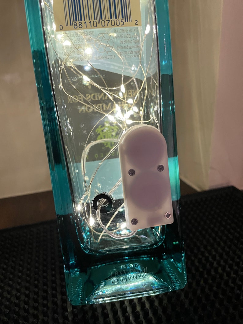 May include: A clear glass bottle with a blue label and a barcode. The bottle is filled with white fairy lights and a small white battery pack. The bottle is sitting on a black surface.