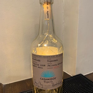 May include: A clear glass bottle of Casamigos Reposado Tequila with a brown label and string lights inside. The label includes text such as "Tequila 100% Agave Azul" and "Jalisco, Mexico". The bottle is sealed with a brown cork.