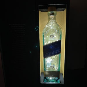 May include: A clear glass bottle with a blue label that says "Johnnie Walker Blue Label" and "Blended Scotch Whisky" inside a black box. The bottle is illuminated with white fairy lights.