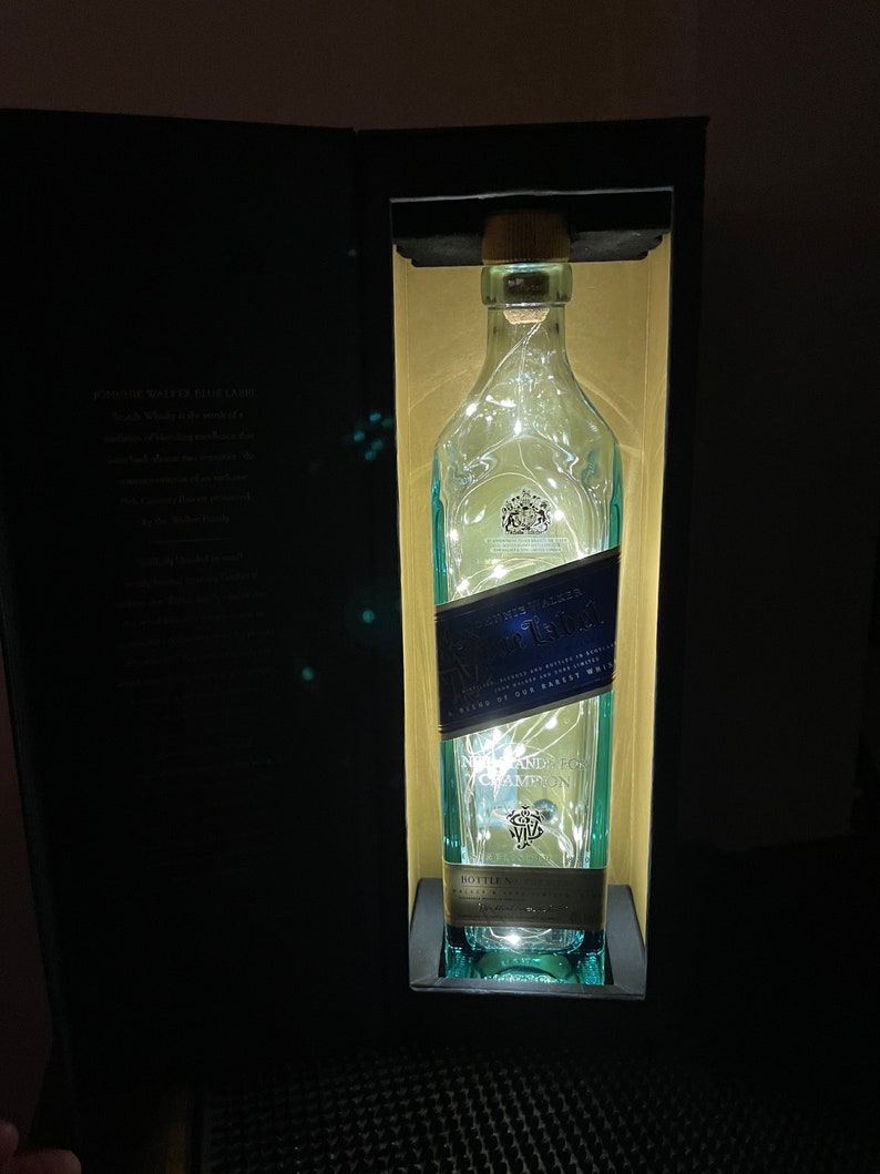 May include: A clear glass bottle with a blue label that says "Johnnie Walker Blue Label" and "Blended Scotch Whisky" inside a black box. The bottle is illuminated with white fairy lights.