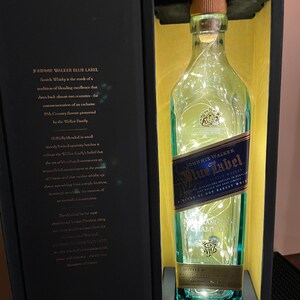 May include: A blue and gold Johnnie Walker Blue Label Scotch Whisky bottle with a white string of lights inside. The bottle is in a black box with a gold interior. The box has text that reads "Scotch Whisky is the result of a tradition of blending excellence that dates back almost two centuries - the commemoration of an exclusive 19th Century flavour pioneered by the Walker Family."