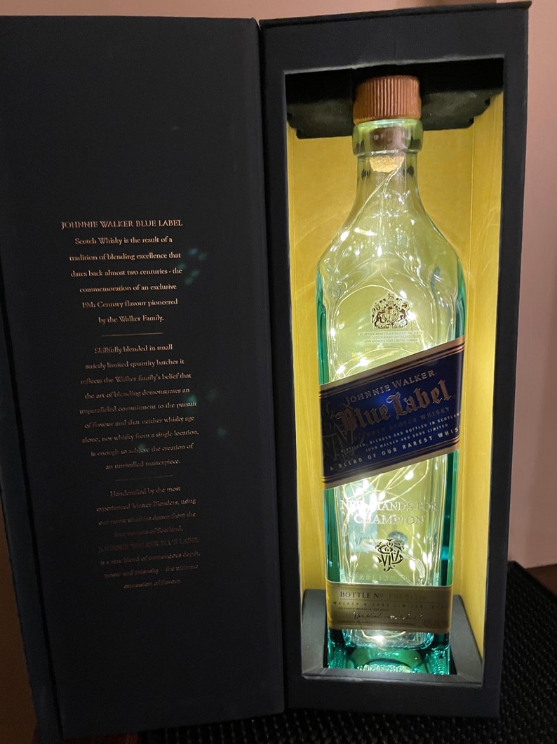 May include: A blue and gold Johnnie Walker Blue Label Scotch Whisky bottle with a white string of lights inside. The bottle is in a black box with a gold interior. The box has text that reads "Scotch Whisky is the result of a tradition of blending excellence that dates back almost two centuries - the commemoration of an exclusive 19th Century flavour pioneered by the Walker Family."