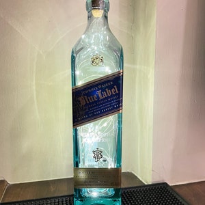 May include: A clear glass bottle with a blue label that reads "JOHNNIE WALKER Blue Label Blended Scotch Whisky A Blend of Our Rarest" and "NED STANDS A CHAMPION". The bottle is decorated with white fairy lights.