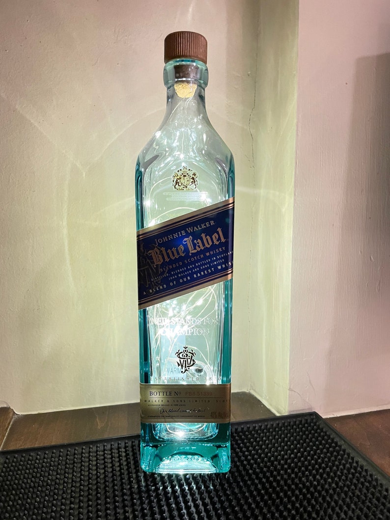 May include: A clear glass bottle with a blue label that reads "JOHNNIE WALKER Blue Label Blended Scotch Whisky A Blend of Our Rarest" and "NED STANDS A CHAMPION". The bottle is decorated with white fairy lights.
