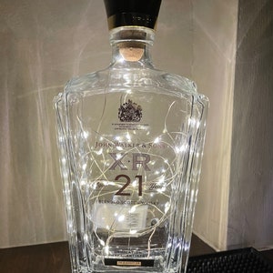 May include: A clear glass bottle with a black cap and a label that reads "John Walker & Sons XR 21 Year Blended Scotch Whisky". The bottle is decorated with white string lights.