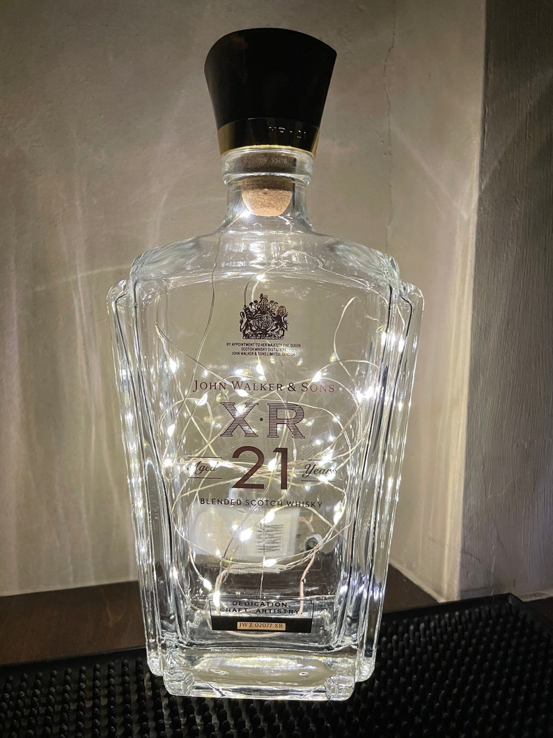 May include: A clear glass bottle with a black cap and a label that reads "John Walker & Sons XR 21 Year Blended Scotch Whisky". The bottle is decorated with white string lights.