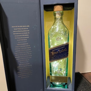 May include: A blue and gold gift box with a clear glass bottle of Johnnie Walker Blue Label Scotch Whisky inside. The bottle is illuminated with white lights. The box has the text "JOHNNIE WALKER BLUE LABEL" and "A BLEND OF OUR RAREST WHISKIES" printed on it.