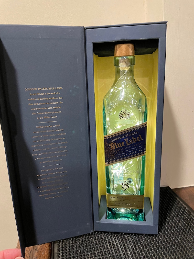 May include: A blue and gold gift box with a clear glass bottle of Johnnie Walker Blue Label Scotch Whisky inside. The bottle is illuminated with white lights. The box has the text "JOHNNIE WALKER BLUE LABEL" and "A BLEND OF OUR RAREST WHISKIES" printed on it.