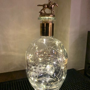 May include: A clear glass bottle with a gold top and a jockey riding a horse figurine. The bottle is decorated with white string lights and has a label that reads "# on Rick No. 103".