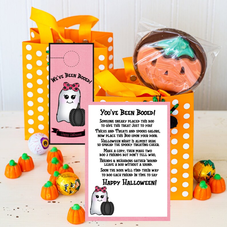 May include: A pink and white printable Halloween "You've Been Booed!" sign with a ghost and pumpkin illustration. The sign includes instructions for a Halloween booing game. The sign is next to a small orange and white polka dot gift bag with a chocolate pumpkin lollipop inside.