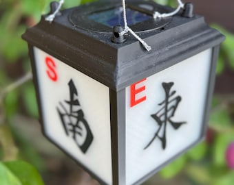 May include: Black and white solar lantern with Chinese characters and the letters "S" and "E" in red. The lantern is hanging from a tree branch. The lantern has a solar panel on top.