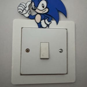 May include: A white light switch cover with a blue Sonic the Hedgehog character sticker on top. Sonic is giving a thumbs up.
