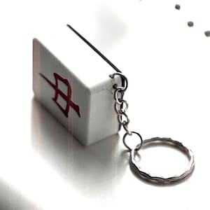 May include: A white rectangular keychain charm featuring a red symbol. It's connected to a silver chain and keyring. The background is white, emphasizing the charm's design and the silver chain.