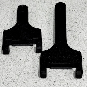 May include: Two black, T-shaped plastic objects. Each has a rectangular base with two short, parallel prongs extending downward. The top portion is a long, flat handle. The objects are on a speckled, light-colored surface.