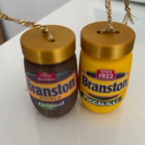 以下が含まれることがあります： 2つのミニチュア瓶のオーナメント。1つは茶色で「Branston Pickle Original」、もう1つは黄色で「Branston Piccalilli」と書かれています。どちらも金色の蓋と吊り下げ用の紐が付いています。