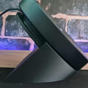 May include: A black speaker with a black stand. The speaker is angled upwards and has a cord plugged into it.