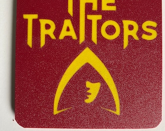 Puede incluir: Un posavasos cuadrado rojo con las palabras "THE TRAITORS" en letras amarillas y un símbolo de aleta de tiburón estilizado amarillo.