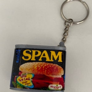 以下が含まれることがあります： スパム缶の形をした灰色のキーホルダー。缶には、黄色い文字で「Spam」と書かれた黒と黄色のラベルが付いています。ラベルには、スパムサンドイッチの写真も載っています。