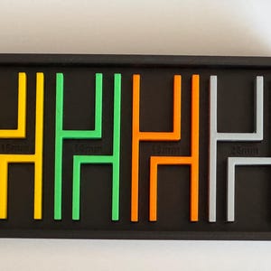 May include: A black tray holds four colorful, plastic letter "H" shapes in yellow, green, orange, and gray. Each letter is labeled with its size in millimeters: 16mm, 14mm, 18mm, and 23mm. The letters are arranged in a row.