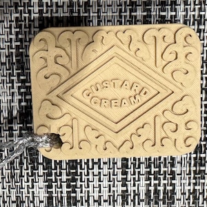 Puede incluir: Una galleta redonda marrón y beige con las palabras "Custard Cream" en relieve en la parte superior. La galleta tiene un patrón decorativo alrededor de los bordes.