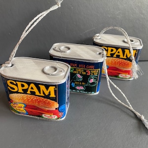 Może przedstawiać: Trzy miniaturowe puszki Spam z niebieskimi i białymi etykietami i czerwonymi i żółtymi akcentami. Każda puszka ma sznurek przymocowany do góry. Puszki znajdują się na szarej powierzchni.