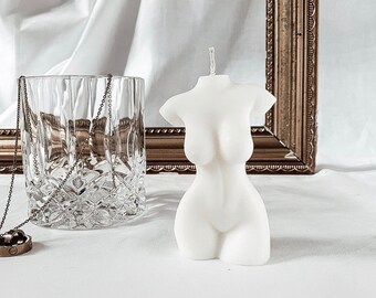 Puede incluir: Vela blanca con forma de torso humano. La vela está sobre una superficie blanca delante de un espejo con marco dorado.