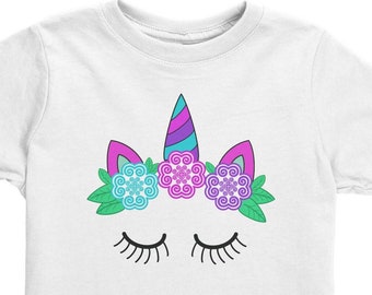 May include: White t-shirt with a unicorn face design. The unicorn has a purple and blue horn, pink ears, and a flower crown with blue, pink, and green flowers. The unicorn has closed eyes with eyelashes.