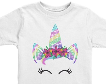 May include: White t-shirt featuring a colorful unicorn design. The unicorn has a rainbow horn, ears, and a floral crown with pink flowers. Below the unicorn design are closed eyelashes.