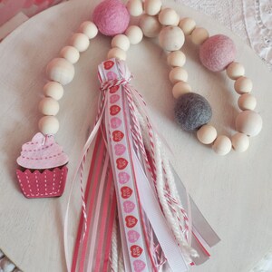 Może przedstawiać: Girlanda walentynkowa z zawieszką w kształcie różowego ciastka z filcu, drewnianymi koralikami i różową i białą wstążką z napisem "Big Hug For Ever".