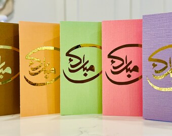 May include: Six greeting cards with a gold foil design of Arabic calligraphy that says "Eid Mubarak". The cards are in different colors: navy blue, brown, orange, green, pink, and purple.