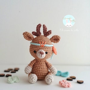 Puede incluir: Un ciervo de crochet marrón con una diadema azul y naranja. El ciervo tiene una cara blanca y patas blancas. El ciervo está sentado sobre una superficie blanca con algunos corazones de madera y otros artículos de crochet.