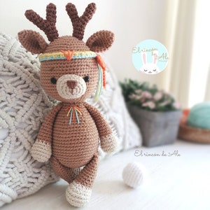 Puede incluir: Un ciervo de crochet marrón con detalles blancos y cuernos. El ciervo lleva una diadema colorida con plumas. El ciervo está de pie sobre una superficie blanca. "El rincon de Ale" está escrito en un círculo blanco con un conejo azul en el centro.