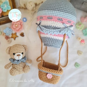 May include: A crocheted hot air balloon with a basket and a crocheted teddy bear. The balloon is grey, pink, and blue with a white stripe. The basket is brown with a pink flower button. The teddy bear is brown with a blue ruffle around its neck.