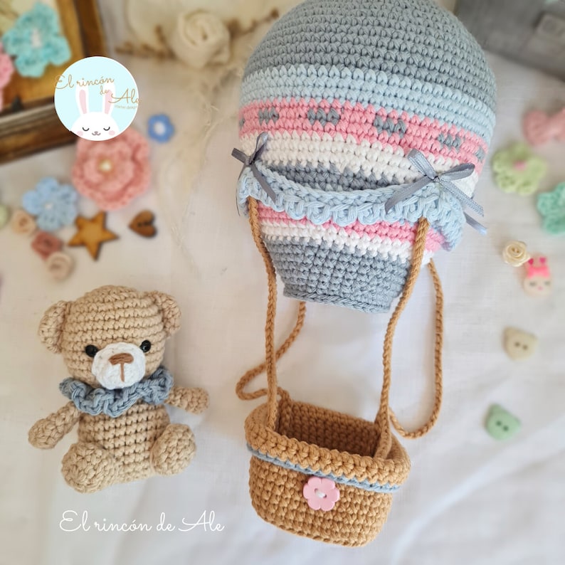 May include: A crocheted hot air balloon with a basket and a crocheted teddy bear. The balloon is grey, pink, and blue with a white stripe. The basket is brown with a pink flower button. The teddy bear is brown with a blue ruffle around its neck.